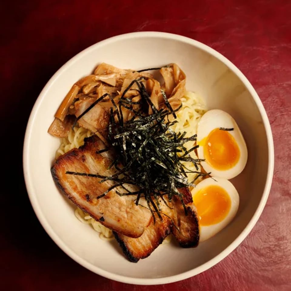 Noodles topped with sliced pork, egg halves, and shredded seaweed on a red table.