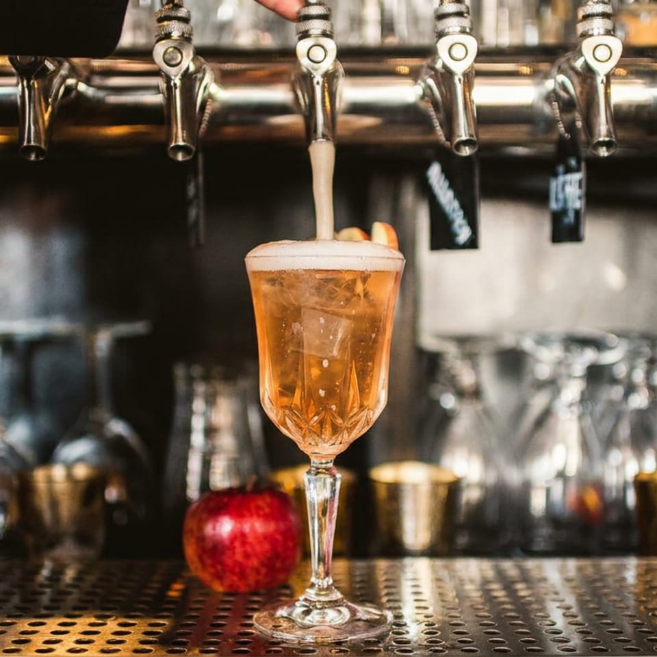 Amber beer pours from a tap into an ornate stemmed glass on a bar, with taps, glassware, and a red apple in the background.
