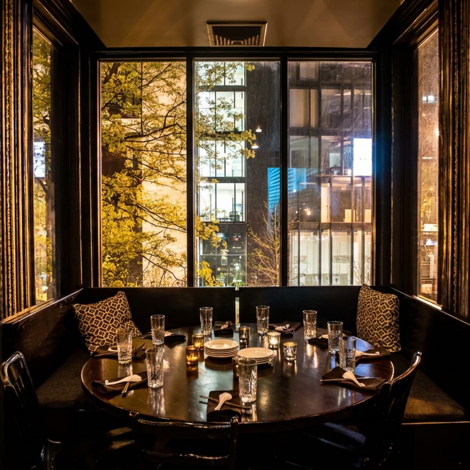 Elegant restaurant booth set with glassware, plates, and candles beside large windows overlooking autumn trees and city buildings.