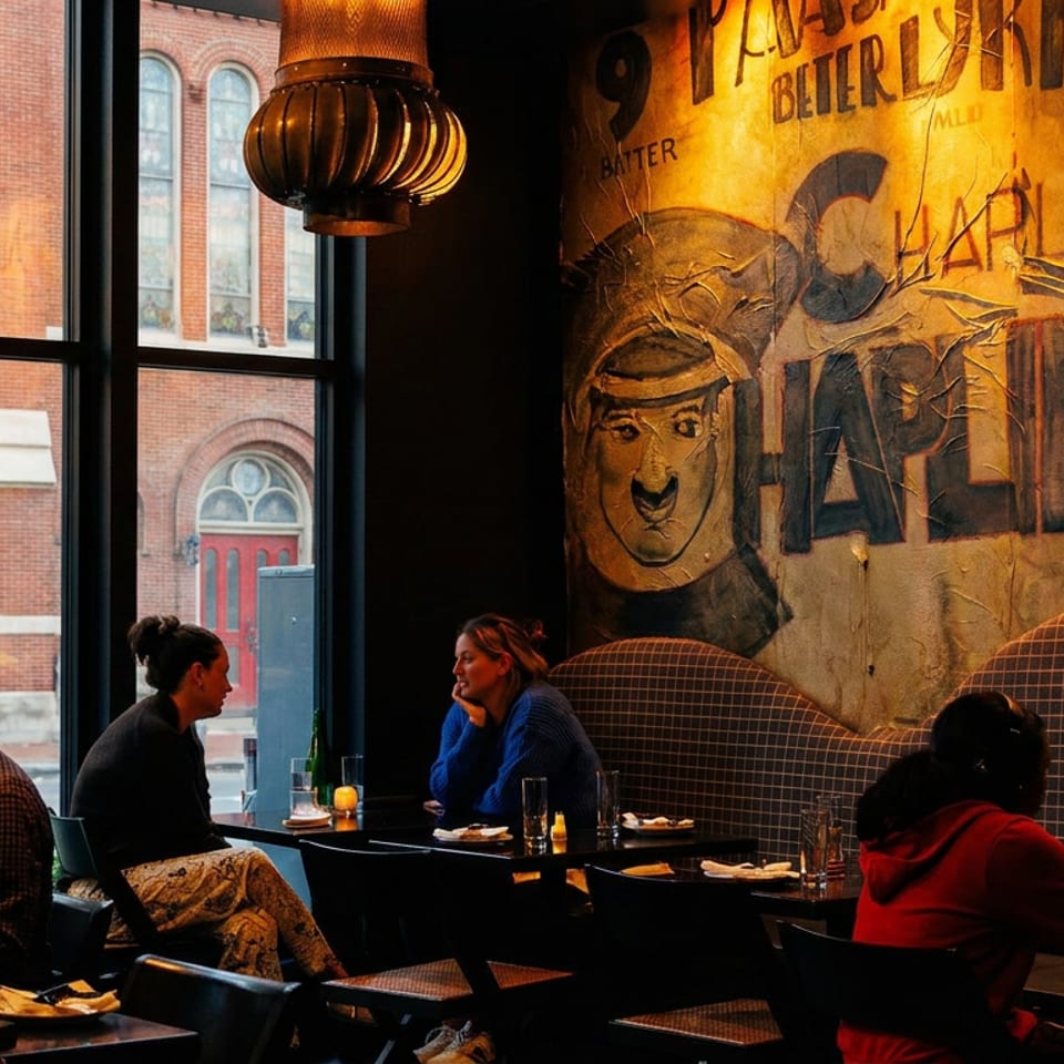 Diners relax at tables inside a warmly lit restaurant with vintage murals, pendant lights, and large windows overlooking a brick building.