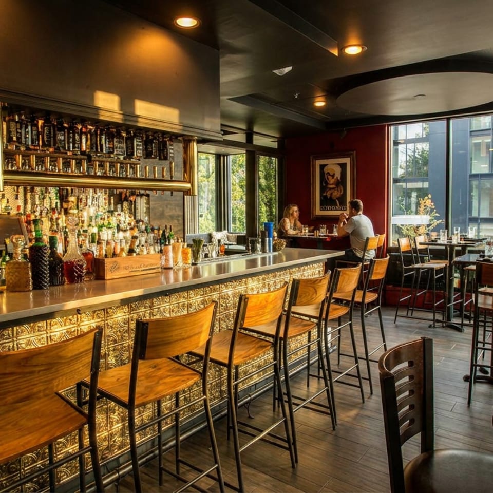 Warmly lit restaurant bar with liquor shelves, bartender, patterned counter, wooden stools, dining tables, and large windows.