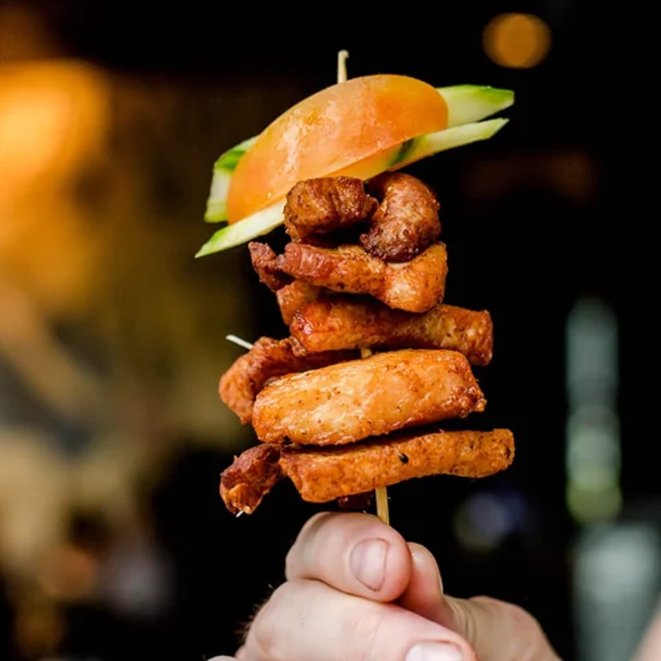 A hand holds a skewer of crispy bites topped with cucumber and tomato.